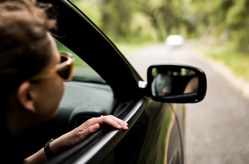 Woman looking out passenger window driving down road with green trees all around.